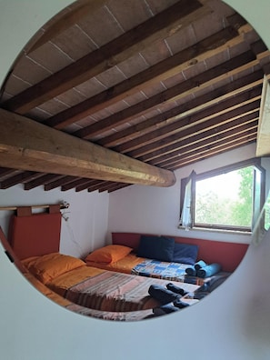 Ein Rundblick auf ein Schlafzimmer mit Holzbalkendecke, einem Fenster und einem Bett mit orange-blauer Bettwäsche.
