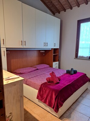 Un dormitorio con cama, armario, ventana y una mesita.