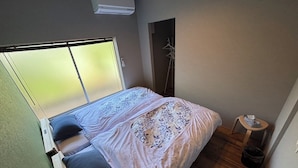 Un dormitorio con cama, un taburete pequeño, una ventana y un equipo de aire acondicionado.
