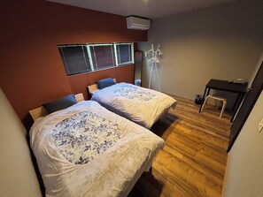 Quarto com duas camas, uma escrivaninha, uma cadeira, janela com persianas e abajur.