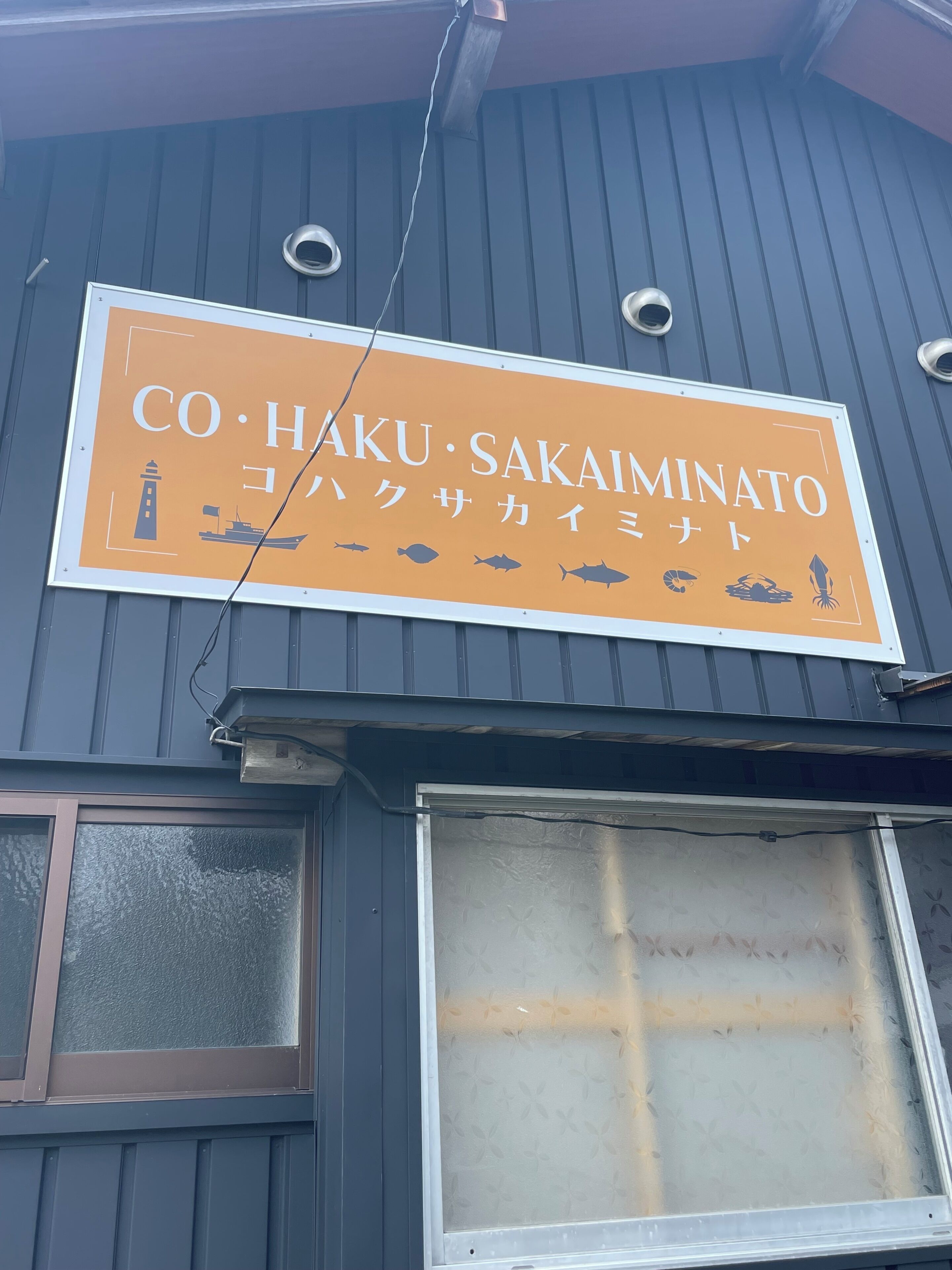 En byggnad med en skylt som visar texten ”CO. HAKU. SAKAINOTTO” och som även innehåller symboler för fisk och ett skepp.