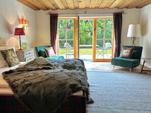 Ein Schlafzimmer mit einem großen Bett, einem grünen Sessel, einer Holzbalkendecke und Blick durch eine Glastür.