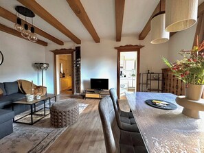 Un salon moderne comprenant un canapé, une table basse, un pouf, une table à manger avec des chaises, un téléviseur et un coin cuisine visible à travers une porte ouverte.