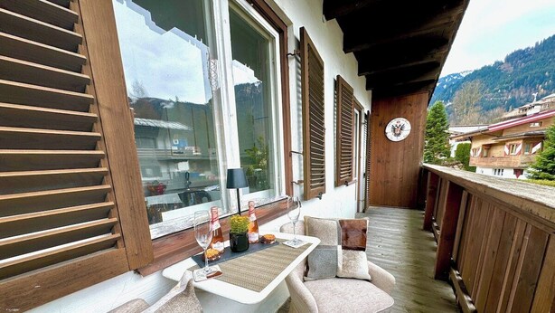 Um deck de madeira com uma mesa, cadeiras, um relógio e vista para edifícios e montanhas.