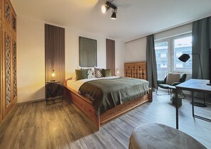 A bedroom with a wooden bed, a small table, a chair, and a window with curtains. - Stylish design apartment for 3-4 people in the heart of Kitzbühel (Kitzbühel)