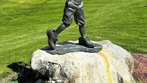 Estátua de uma pessoa com um taco de beisebol em uma rocha, com um campo de golfe e montanhas ao fundo.