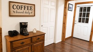 Un mueble de madera con una cafetera y tazas, una puerta con un letrero que dice 'COFFEE preparado a diario', y un pasillo con piso de madera y una puerta blanca.