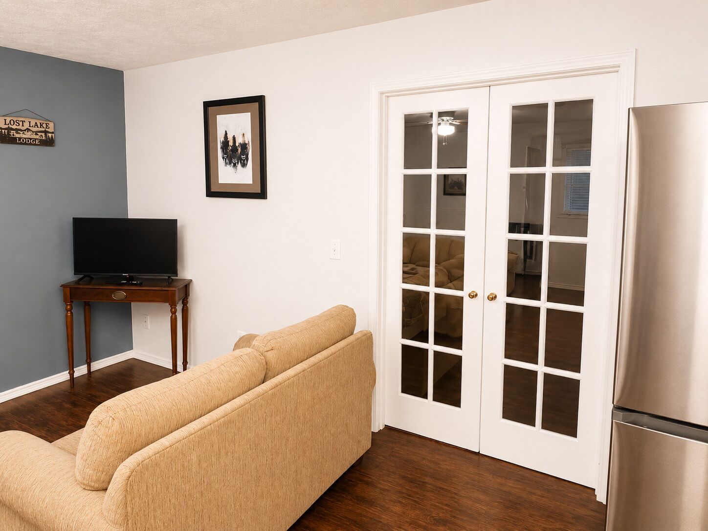 A living room with a beige sofa, a television on a wooden table, a framed picture on the wall, and a white door with glass panels.