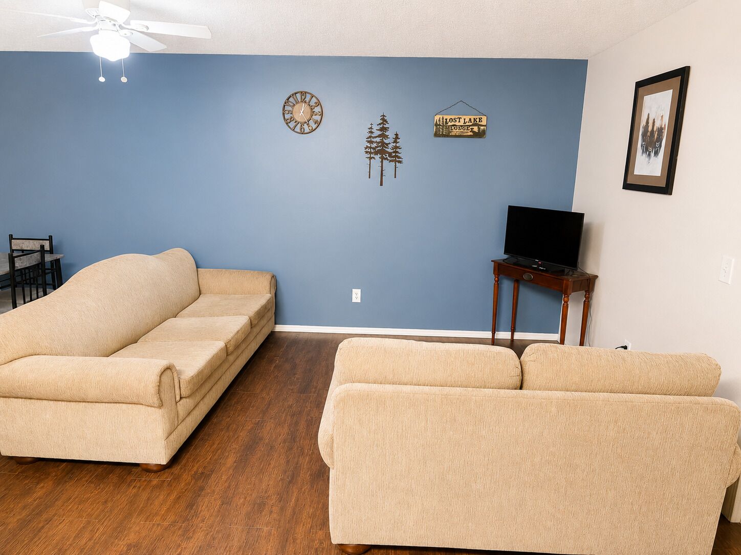 Ein Wohnzimmer mit blauer Wand, zwei beigefarbenen Sofas, einem Holztisch, einem Deckenventilator, einer Uhr, einem Schild mit der Aufschrift 'Lost Lake Lodge' und einem gerahmten Bild an der Wand.
