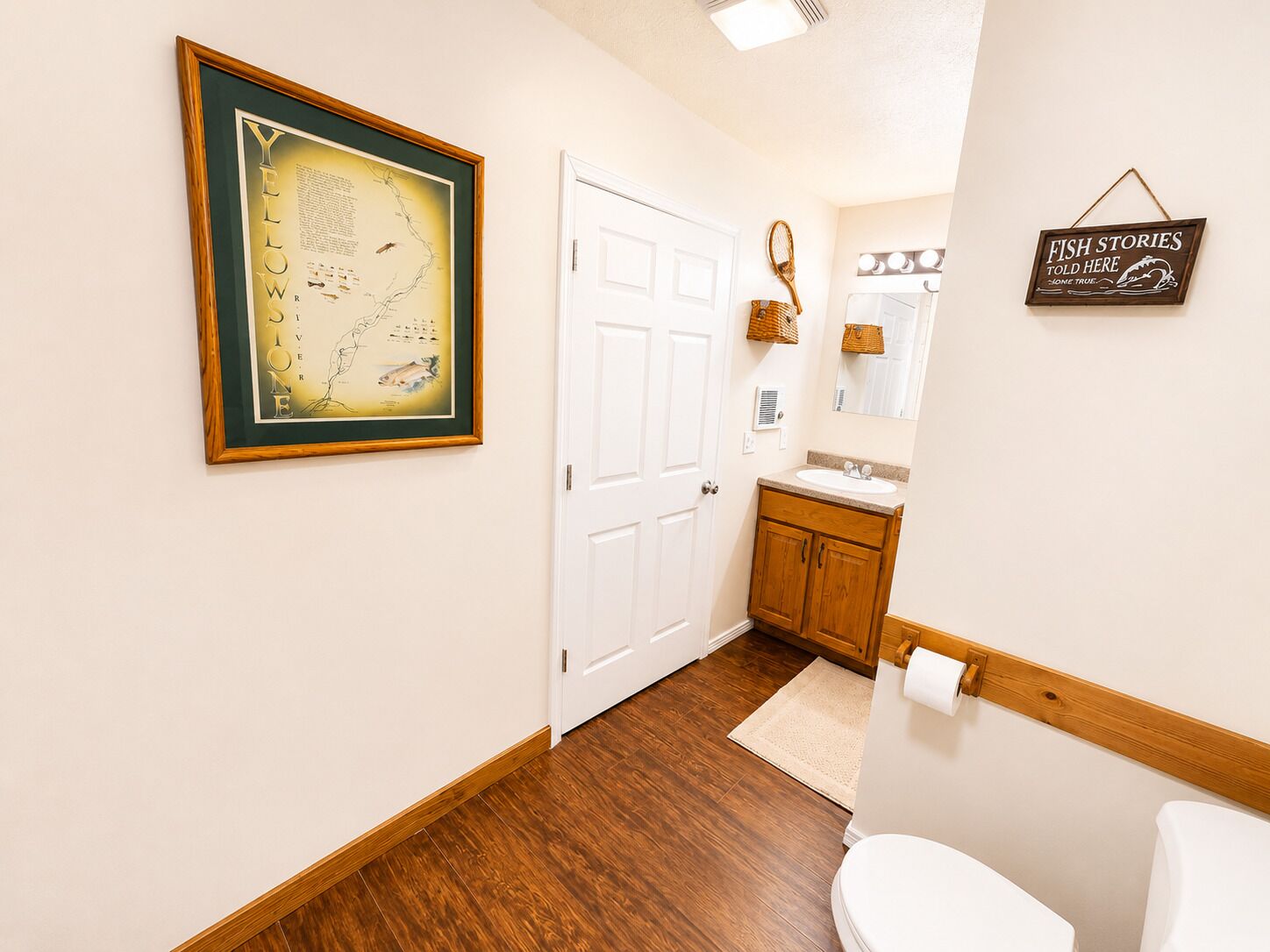 Un couloir avec une carte encadrée, une porte blanche, un plancher en bois et une salle de bain équipée d’un lavabo, d’un miroir et d’une pancarte.