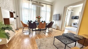 Een moderne woonkamer met een eetgedeelte, houten vloeren, een groot raam met gordijnen, een flatscreen-tv en een kamerplant.