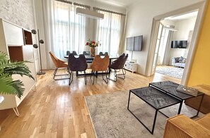 Ein modernes Wohnzimmer mit Essbereich, Holzboden, einem großen Fenster mit Vorhängen, einem Flachbildfernseher und einer Topfpflanze.
