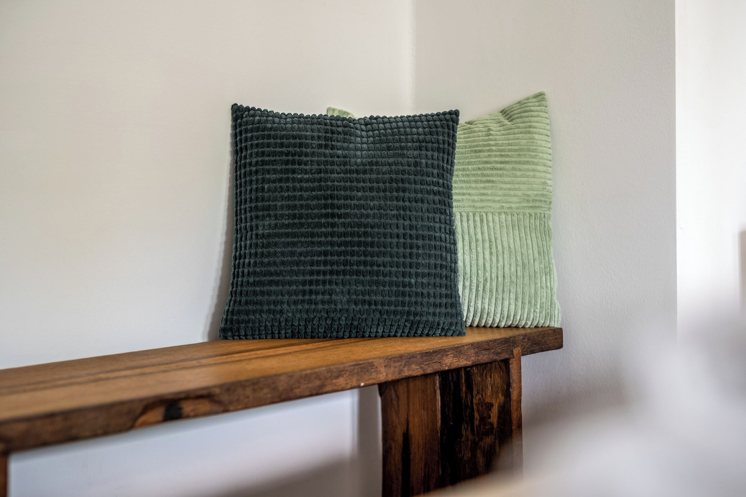 Two pillows on a wooden bench against a plain wall.