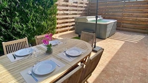 Une terrasse extérieure avec une table en bois dressée pour les repas, un jacuzzi et un grand parasol offrant de l’ombre.