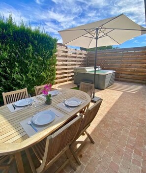 An outdoor patio with a wooden table set for dining, a hot tub, and a large umbrella providing shade. - Renovated 80-square-meter Retreat with Spa & Enclosed Garden - Near Futuroscope (Beaumont Saint-Cyr)