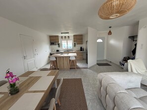 En moderne stue med en sofa, spisebord med stole, et køkken med træskabe og en hængende lampe.