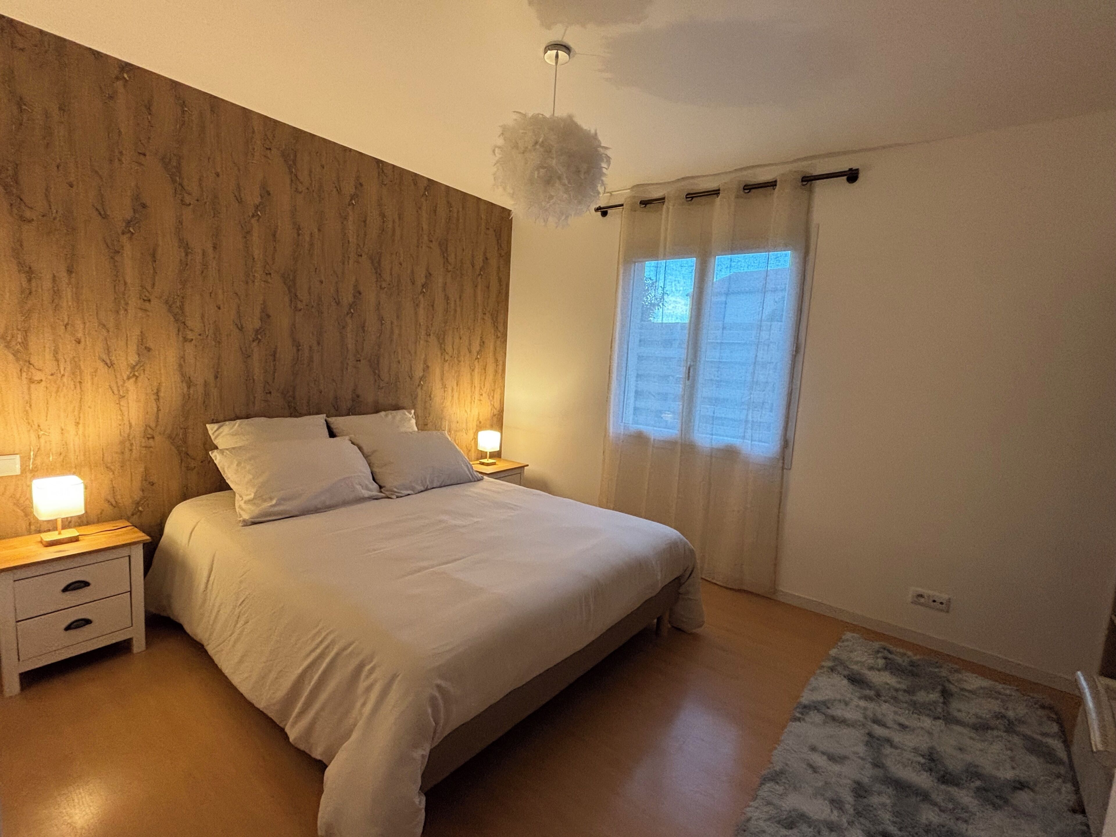A bedroom with a wooden headboard, a bed with white bedding, two bedside tables with lamps, and a window with curtains.