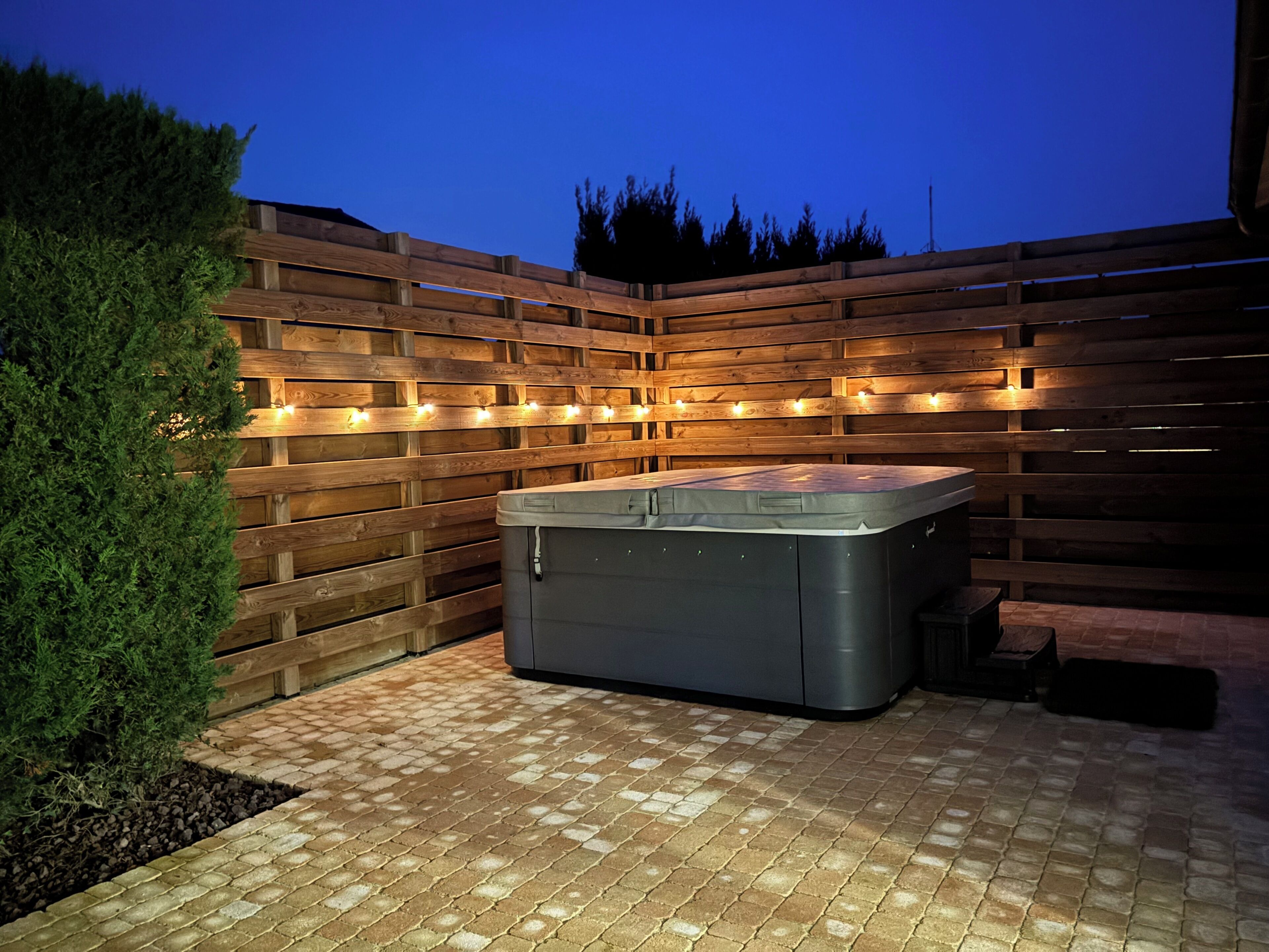 Un jacuzzi en un patio exterior con un cerramiento de madera y luces colgantes.