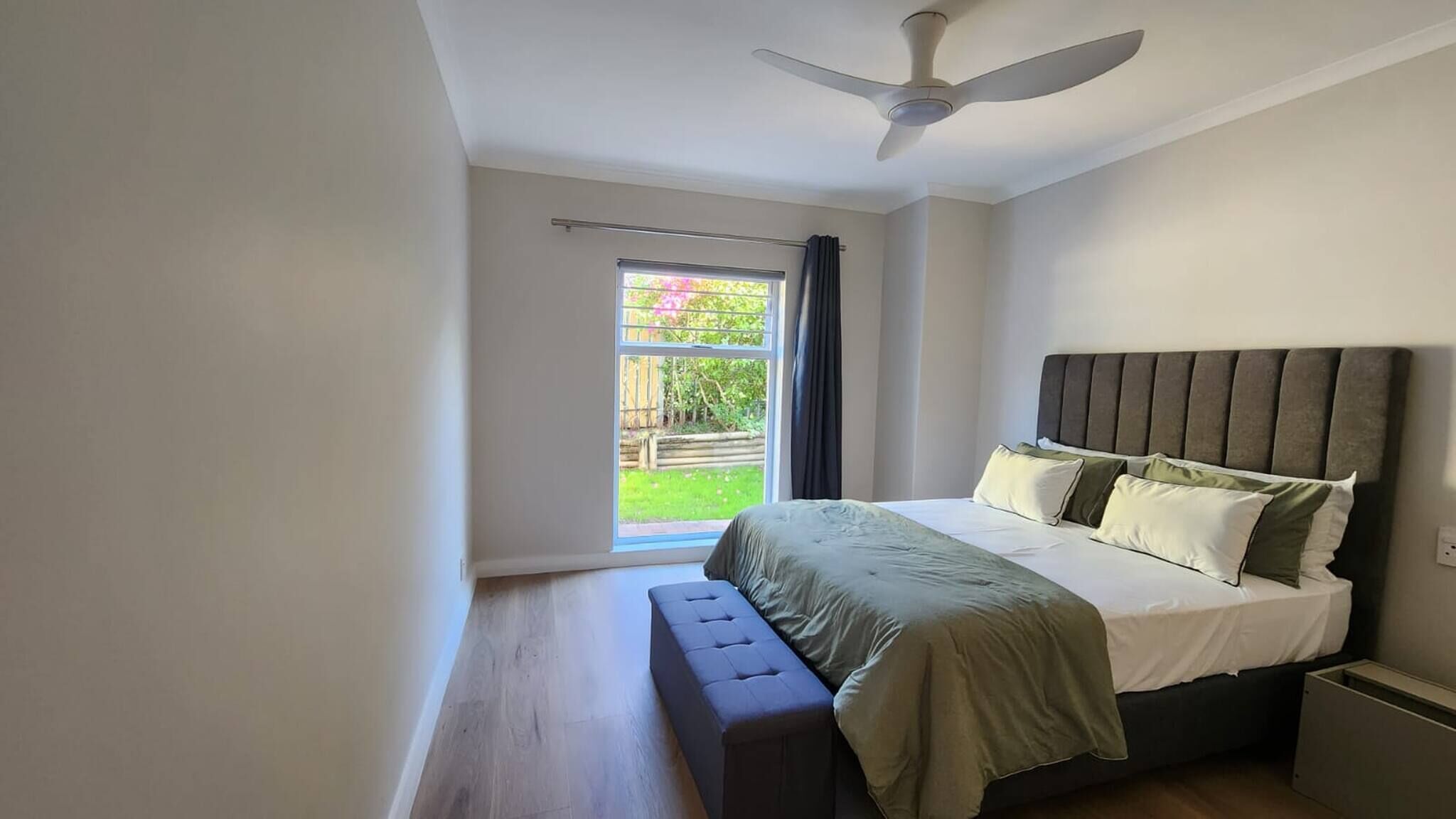 A bedroom with a bed, a bench, a ceiling fan, and a view of a garden through a window.
