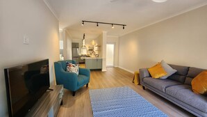 Una sala de estar con televisión, sofá y sillón, una cocina al fondo y una alfombra estampada.