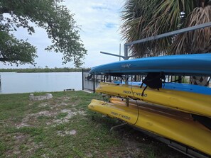 Due kayak, uno blu e uno giallo, ormeggiati vicino a un corso d'acqua, con una recinzione e alberi sullo sfondo.