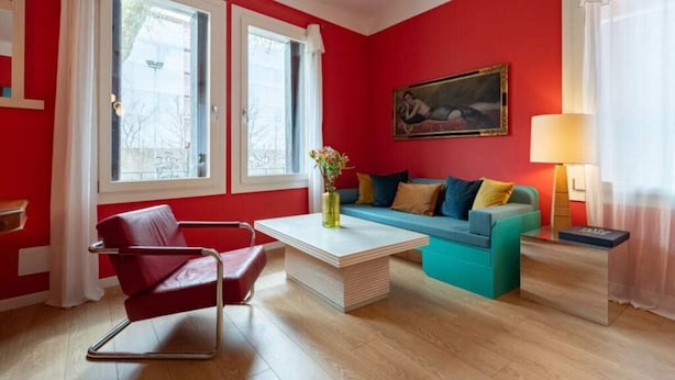 A room with a red wall, a sofa, a chair, a coffee table, a painting, and a lamp. - Charming Venetian  Retreat-3 Bedrooms, Dorsoduro (Venezia)