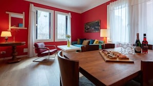 A red-walled room with a wooden dining table, two windows with white curtains, a sofa, a chair, and a lamp. - Charming Venetian  Retreat-3 Bedrooms, Dorsoduro (Venezia)