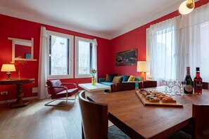 A red-walled room with a wooden dining table, two windows with white curtains, a sofa, a chair, and a lamp. - Charming Venetian  Retreat-3 Bedrooms, Dorsoduro (Venezia)