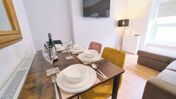 Zona pranzo con tavolo apparecchiato per quattro persone, televisore a parete e divano sullo sfondo.
