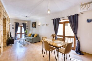 Ein Wohnzimmer mit Sofa, einem Esstisch mit Stühlen, einem Fernseher und einem Fenster mit Vorhängen.