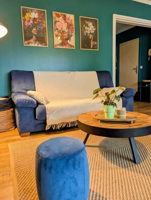 Un salon avec un canapé bleu, une table basse ronde ornée d’une plante et trois tableaux aux motifs floraux accrochés au mur.