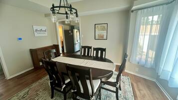 Uma sala de jantar com mesa e cadeiras de madeira, um candelabro e uma cozinha ao fundo.