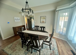 A dining room with a wooden table and chairs, a chandelier, and a kitchen in the background.