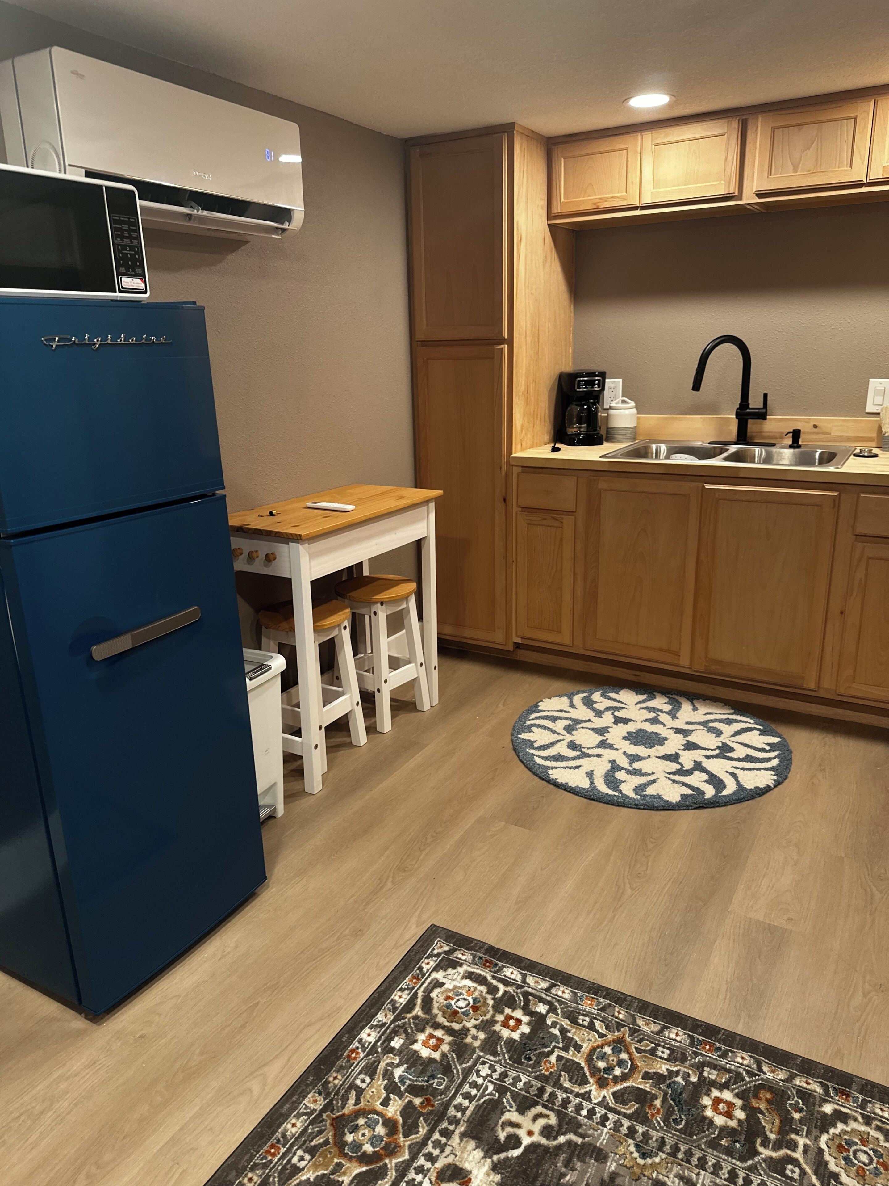A compact kitchen with a blue refrigerator, wooden cabinets, a sink, and a round rug.