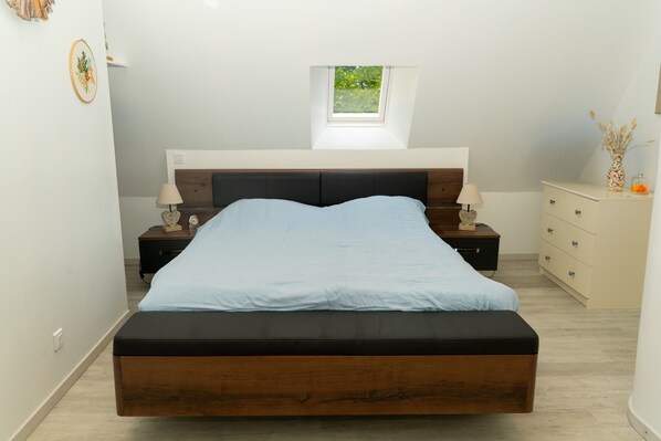 Une chambre à coucher comprenant un lit, des tables de chevet, une commode, une fenêtre et une petite lucarne.