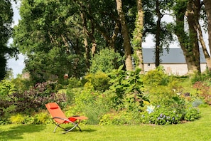 A garden with a red chair, lush greenery, and a building in the background. - Guest Room "Suite Le Bocage" (Pleine-Sève)