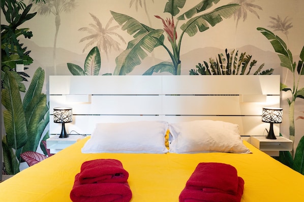 A bed with a yellow bedspread, white pillows, and red towels. There are two bedside lamps with black bases and white shades. The headboard is white with horizontal lines. The wall behind the bed features a tropical mural with palm trees and large leaves.