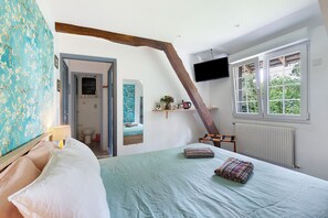 Un dormitorio con cama, televisor montado en la pared, ventana con vista a follaje y puerta que da a otra habitación.