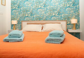 Un lit bien fait, recouvert d’une couvre-lit orange, avec deux serviettes bleues de chaque côté, et un papier peint à motifs floraux en arrière-plan.