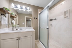 A bathroom with a glass shower enclosure, a vanity with a mirror, and a shelf with plants. - 3-Bedroom Townhome - The Gardens Cottage Duo Unit A (Houston)