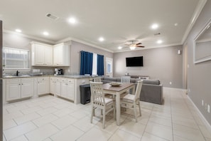 A modern kitchen with white cabinets, a dining table with chairs, a gray sofa, and a ceiling fan. - 3-Bedroom Townhome - The Gardens Cottage Duo Unit A (Houston)