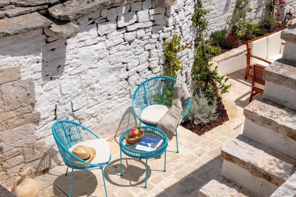 A stone wall, two turquoise chairs with cushions, a small table with a book and a hat, and a staircase.