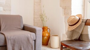 Een kamer met een grijze bank, een houten stoel, een vaas met gedroogde planten en een strohoed op de stoel.