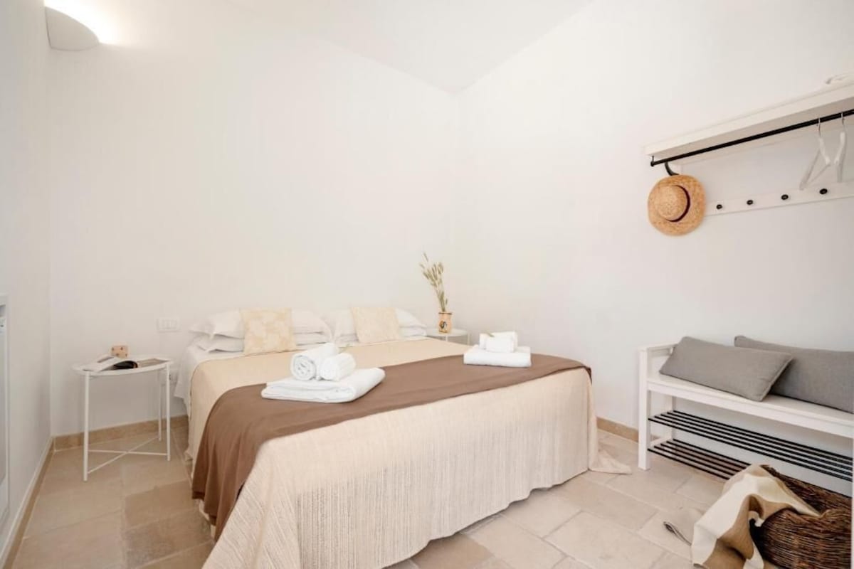 Un dormitorio con cama, una mesita de noche, un banco y un sombrero en la pared.