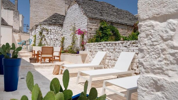 Een gebouw van steen met een witte gevel, een terras met witte loungestoelen en potplanten.