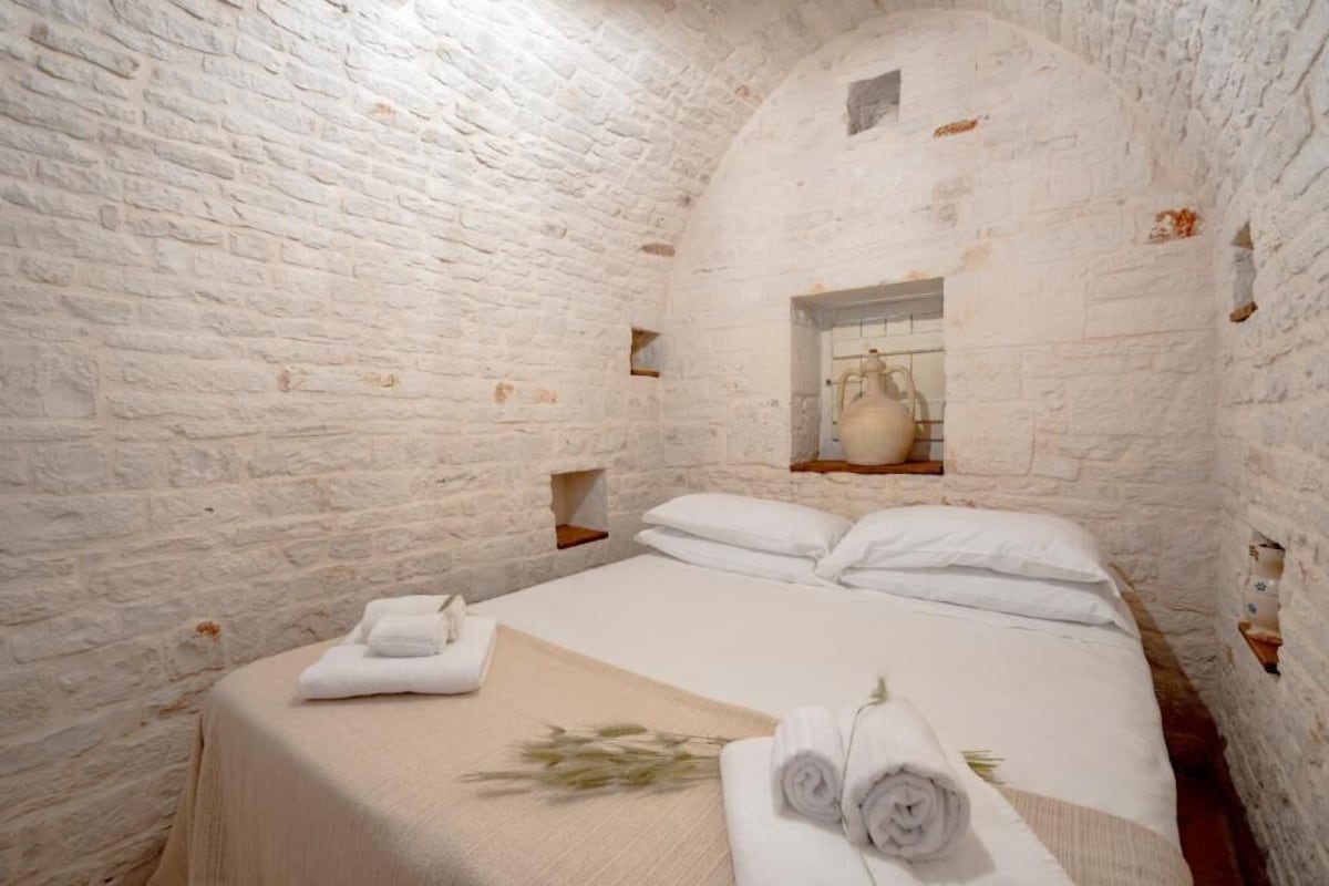 Una habitación con una cama, ropa de cama blanca, una ventana con un jarrón y una pared de ladrillo.