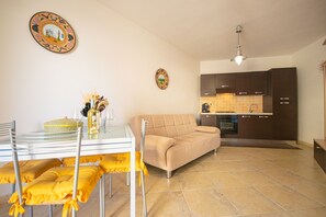 A modern kitchen with dark cabinets, a dining area with a glass table and yellow cushions, and a beige sofa. - Casa delle Sorgenti 1 (Torpè)
