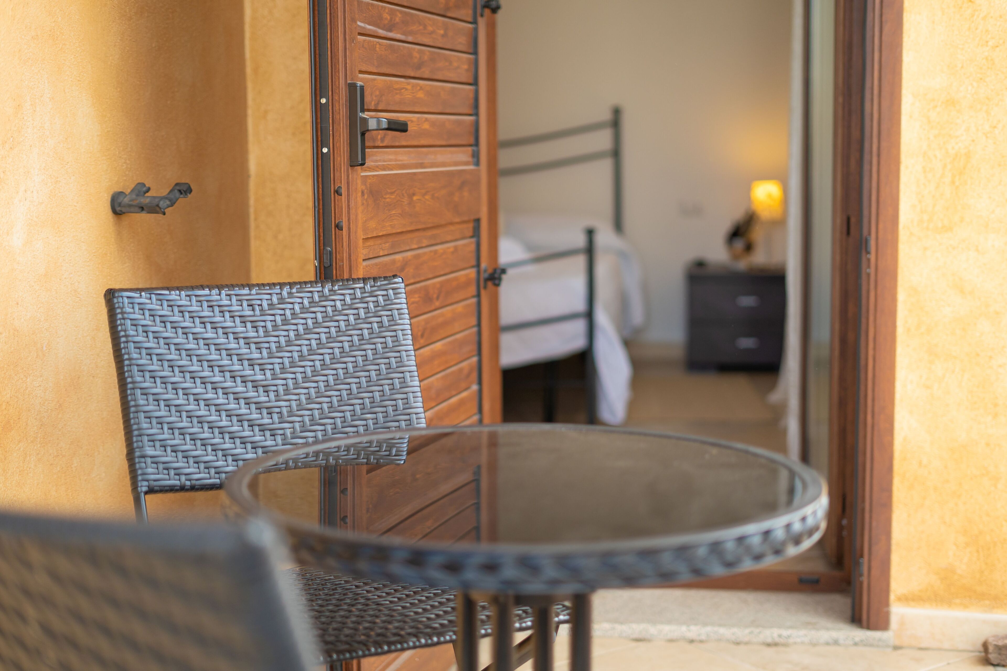 A room with a wicker chair, a glass table, a wooden door, and a bed with a nightstand.