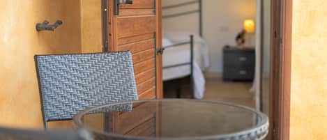 A room with a wicker chair, a glass table, a wooden door, and a bed with a nightstand.