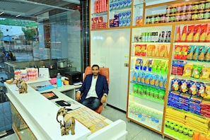 A man seated in an office chair at a reception desk with various packaged goods and a refrigerator stocked with beverages.
